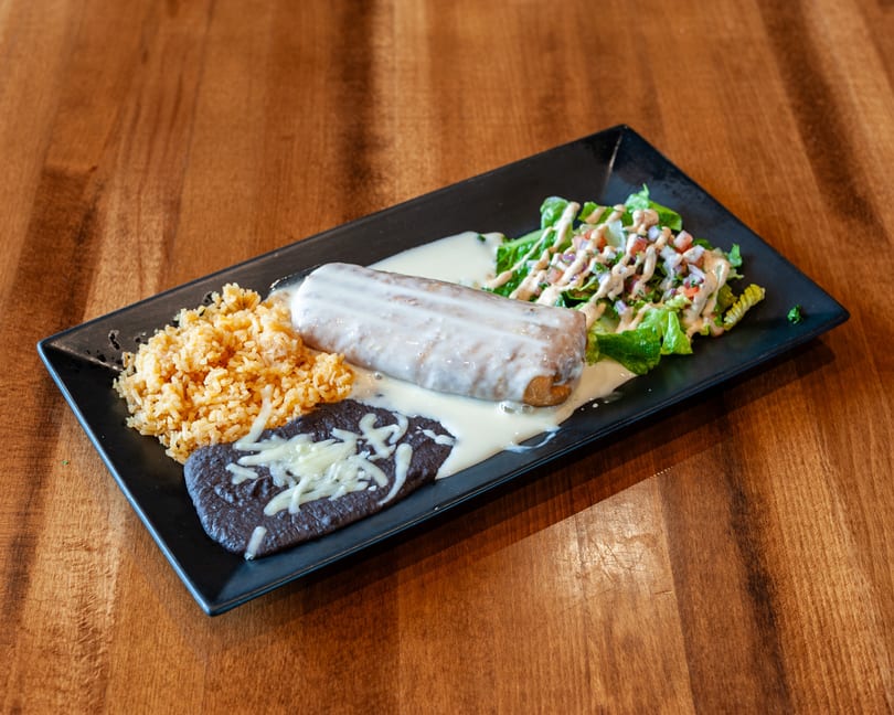 A burrito covered in white sauce, with rice, refried beans, and salad on a black plate.