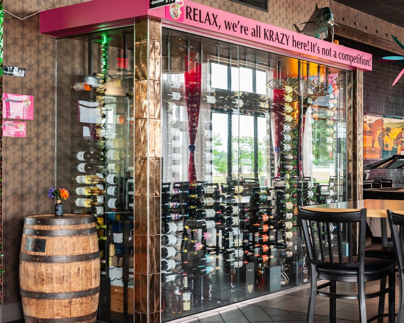 A glass-enclosed wine cellar with a pink sign reading "RELAX, we're all KRAZY here! It's not a competition!"