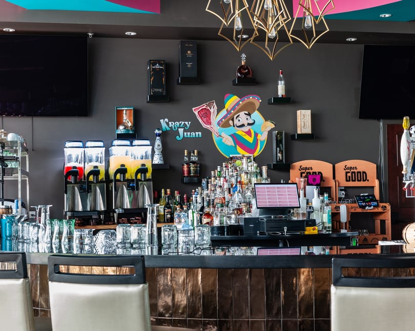 A vibrant bar with a "Krazy Juan" logo, margarita machines, liquor bottles, and bar stools.