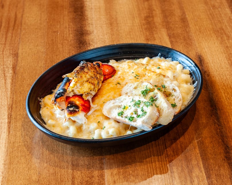 Lobster mac and cheese with toasted bread slices and parsley in a black bowl on a wooden table.
