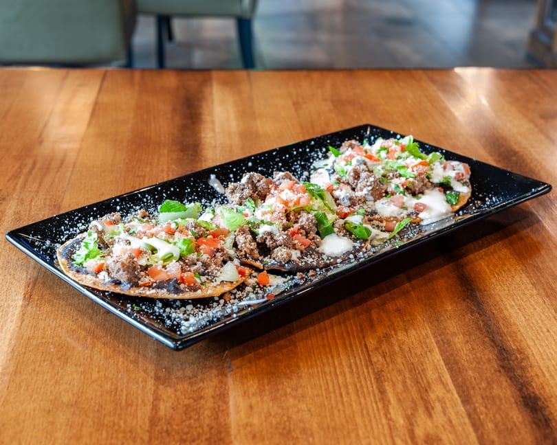 Three tostadas with ground beef, lettuce, tomatoes, and sour cream on a black rectangular plate.