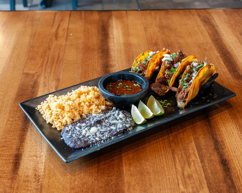 Four birria tacos with rice, refried beans, limes, and a dipping sauce on a black plate.
