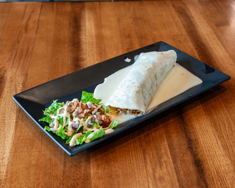A large burrito covered in white sauce, served with a side salad on a black rectangular plate.
