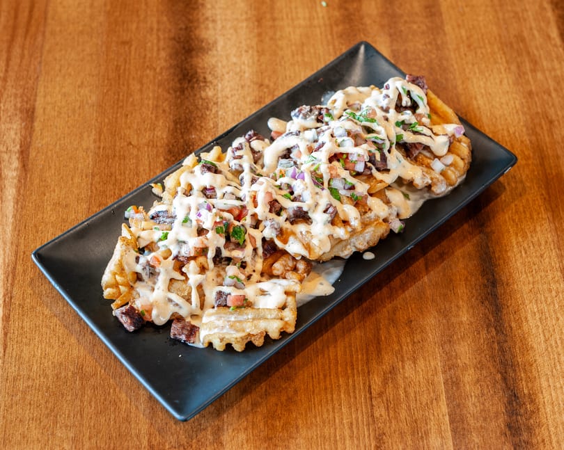 Waffle fries topped with steak, pico de gallo, and a creamy white sauce on a black plate.