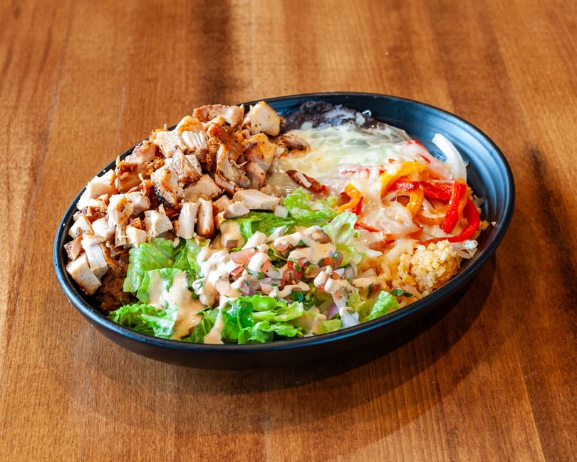 A colorful chicken bowl with lettuce, pico de gallo, rice, beans, cheese, and peppers.