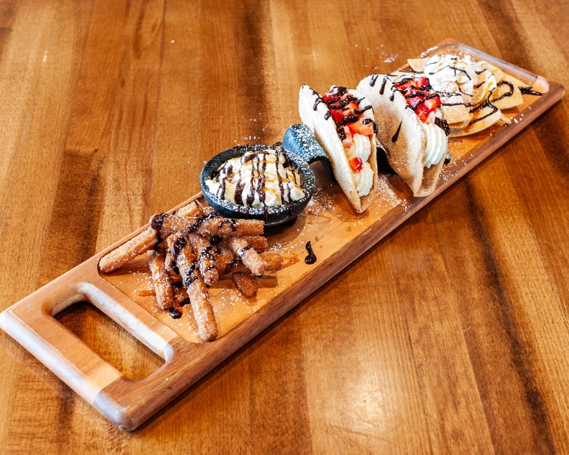 Dessert platter with churros, ice cream, dessert tacos, and mini pancakes on a wooden board.