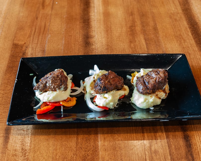 Three portions of meat and mashed potatoes with peppers and onions on a black rectangular plate.