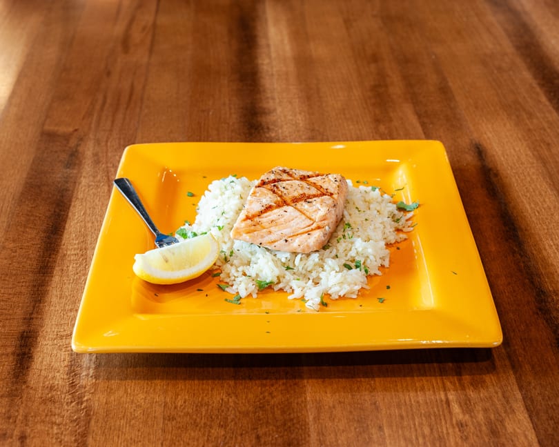 Grilled salmon on a bed of white rice with parsley, a lemon wedge, and a fork on a yellow plate.